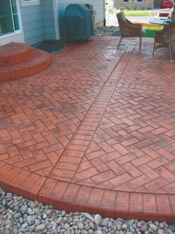 A rustic red concrete backyard patio is colored by dry granular pigments and stamped with an alternating brick pattern