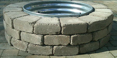 Image of a round firepit/fireplace made of tan colored bedford reading rock stone, with a metal ring sitting in the middle.